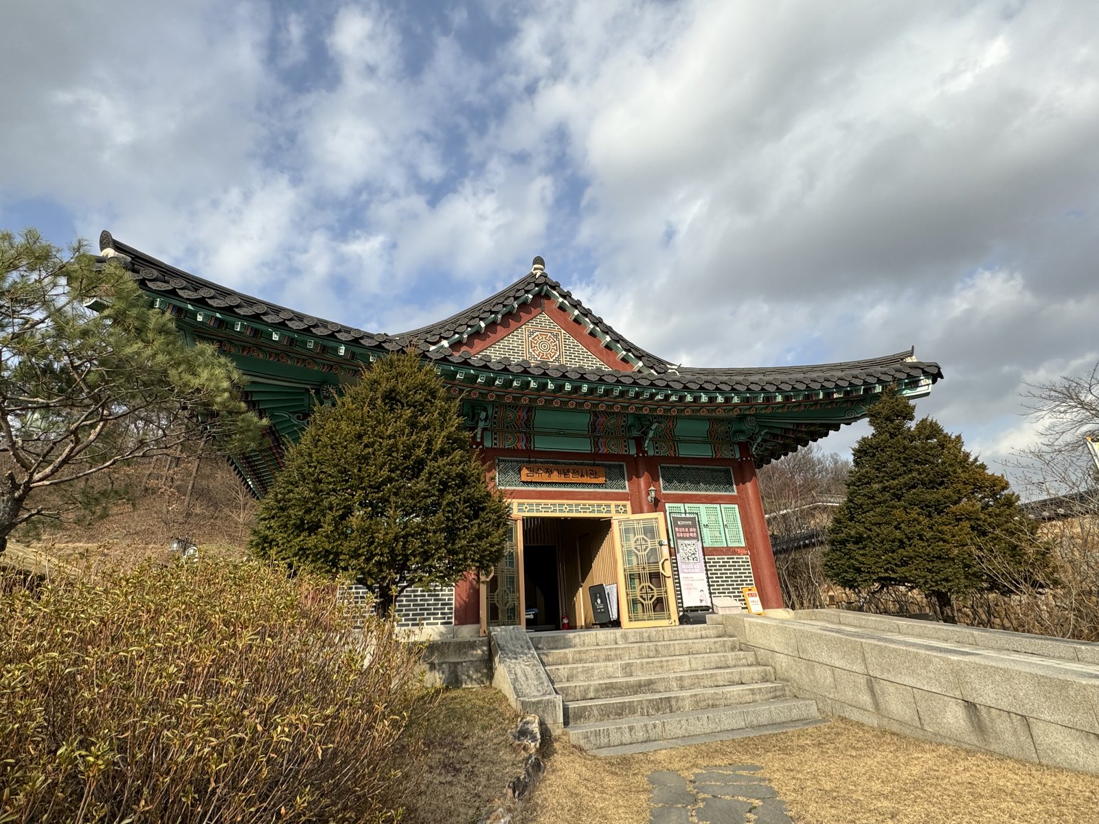 구름 낀 하늘 아래 단청을 칠한 기와 건물을 올려다보았다. 문 위 나무 현판에 김유정기념전시관이라고 쓰여 있고, 앞에 돌계단과 양옆 향나무가 있다.