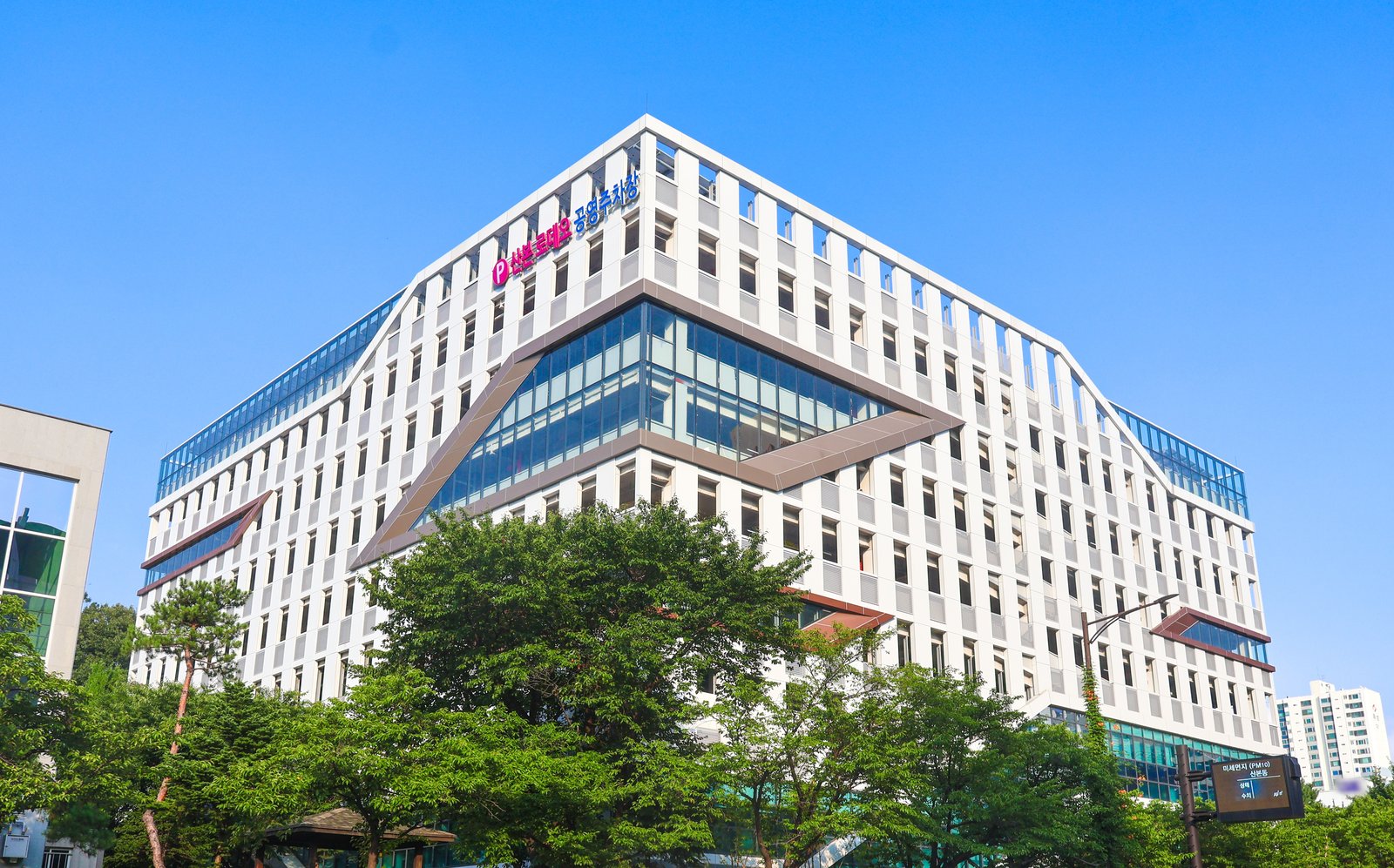흰 틀이 촘촘한 큰 건물 위쪽 모서리에 산본로데오 공영주차장이라는 글씨가 있다. 건물 앞에 초록 나무가 우거지고, 오른쪽에 미세먼지 알림판이 서 있다.