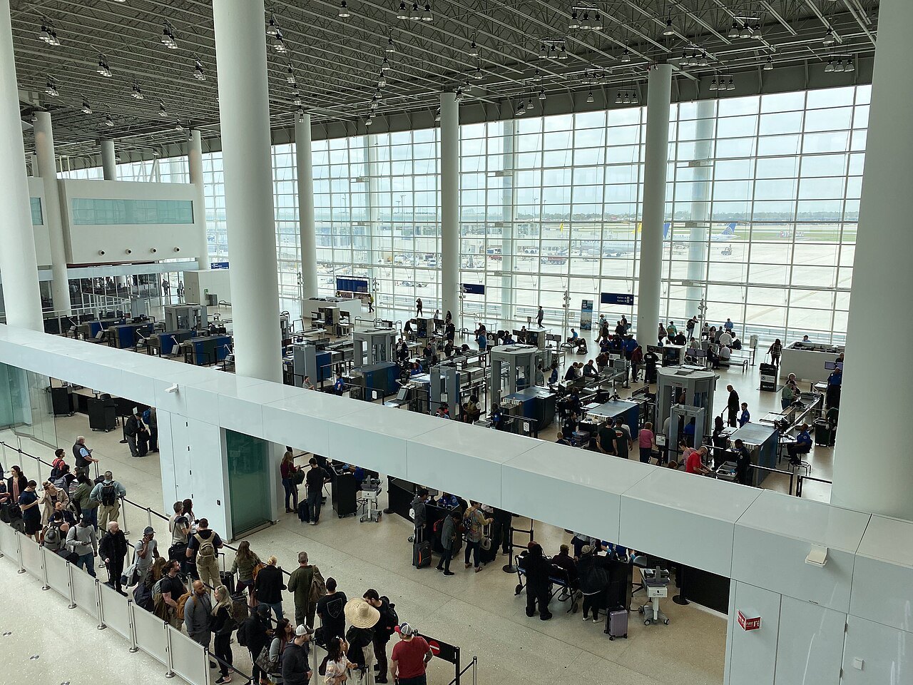 위층에서 내려다본 공항 보안 검색장 — 줄 선 승객들과 여러 줄의 검색 장비, 통과 문형 탐지기, 유리창 너머 활주로의 비행기