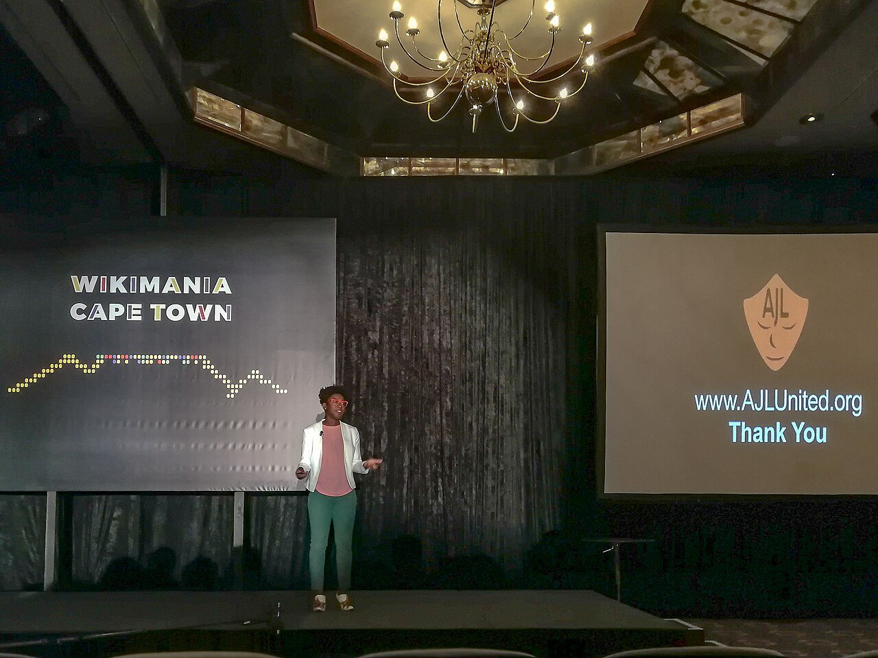 어두운 강당 무대에서 발표하는 조이 부올람위니. 왼쪽 화면에는 WIKIMANIA CAPE TOWN, 오른쪽 화면에는 AJL 가면 로고와 www.AJLUnited.org Thank You가 떠 있다