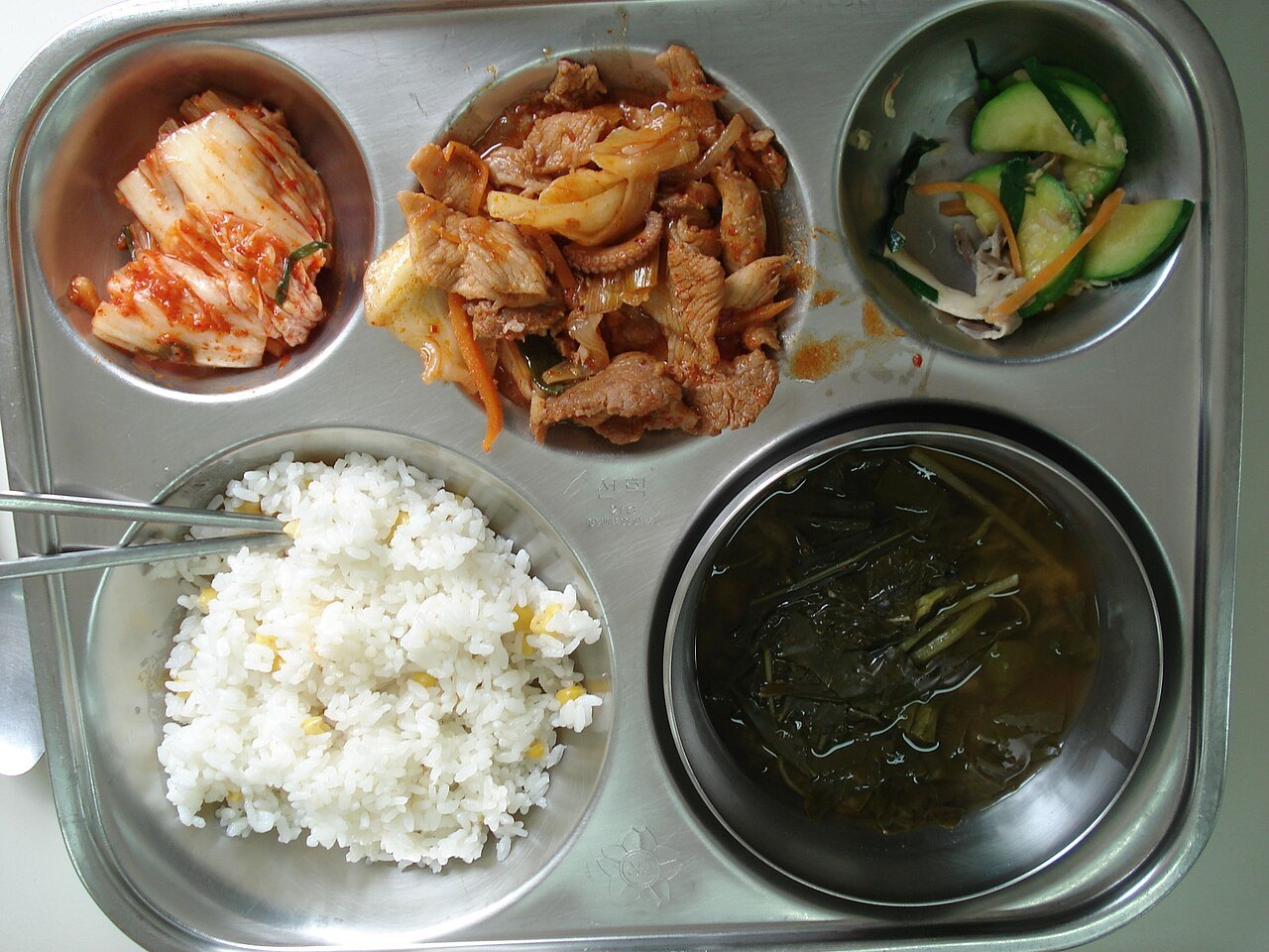 칸이 나뉜 스테인리스 식판에 담긴 한국 학교 급식 — 배추김치, 양파를 넣은 매콤한 고기 볶음, 채소 무침, 옥수수를 섞은 밥, 잎채소가 든 국