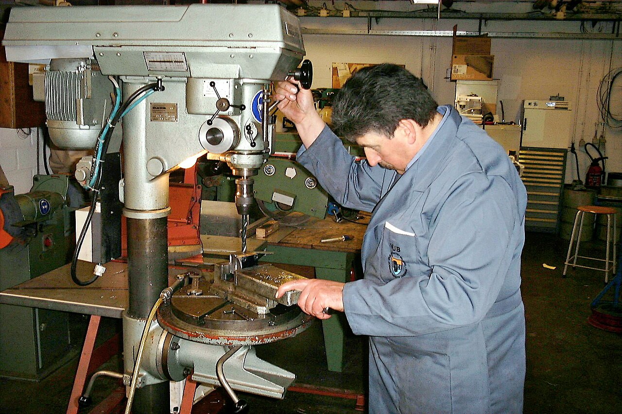 작업복을 입은 사람이 작업장에서 탁상 드릴의 손잡이를 내려 고정된 금속 부품에 구멍을 뚫는 모습